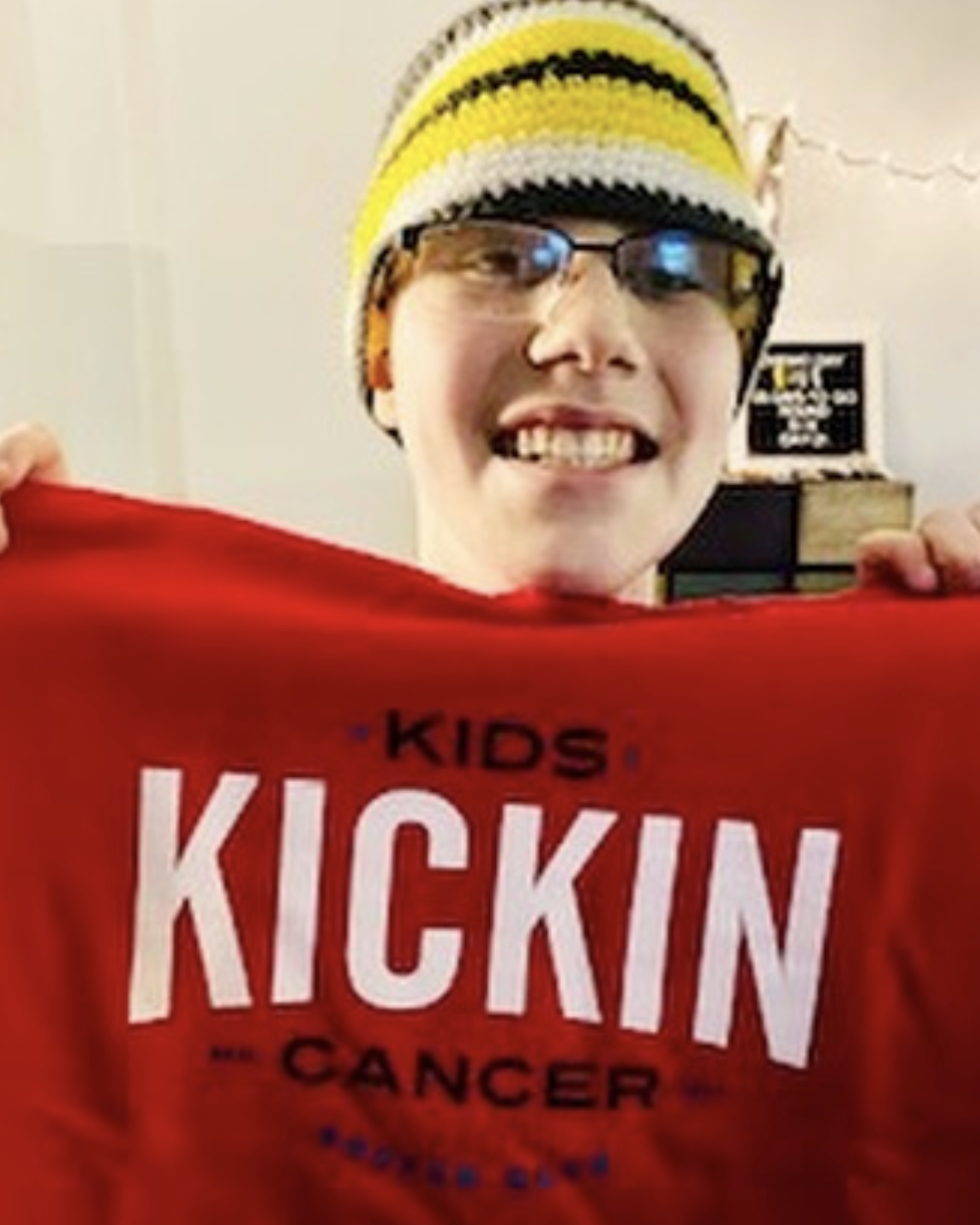 Cancer Kickers Teammate Jay wearing a hat smiling and holding up red t-shirt with cancer kickers logo nike label