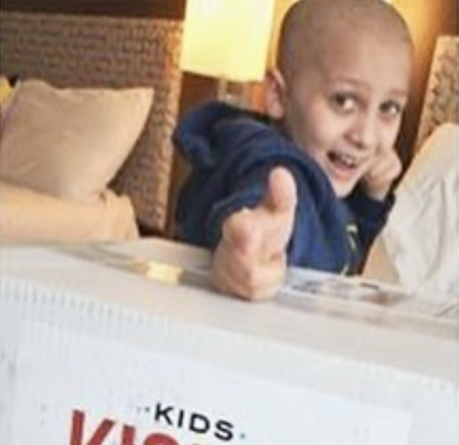 Cancer Kickers Teammate Jude wearing player kit gear blue hoodie giving a thumbs up and smiling over shipping box with logo